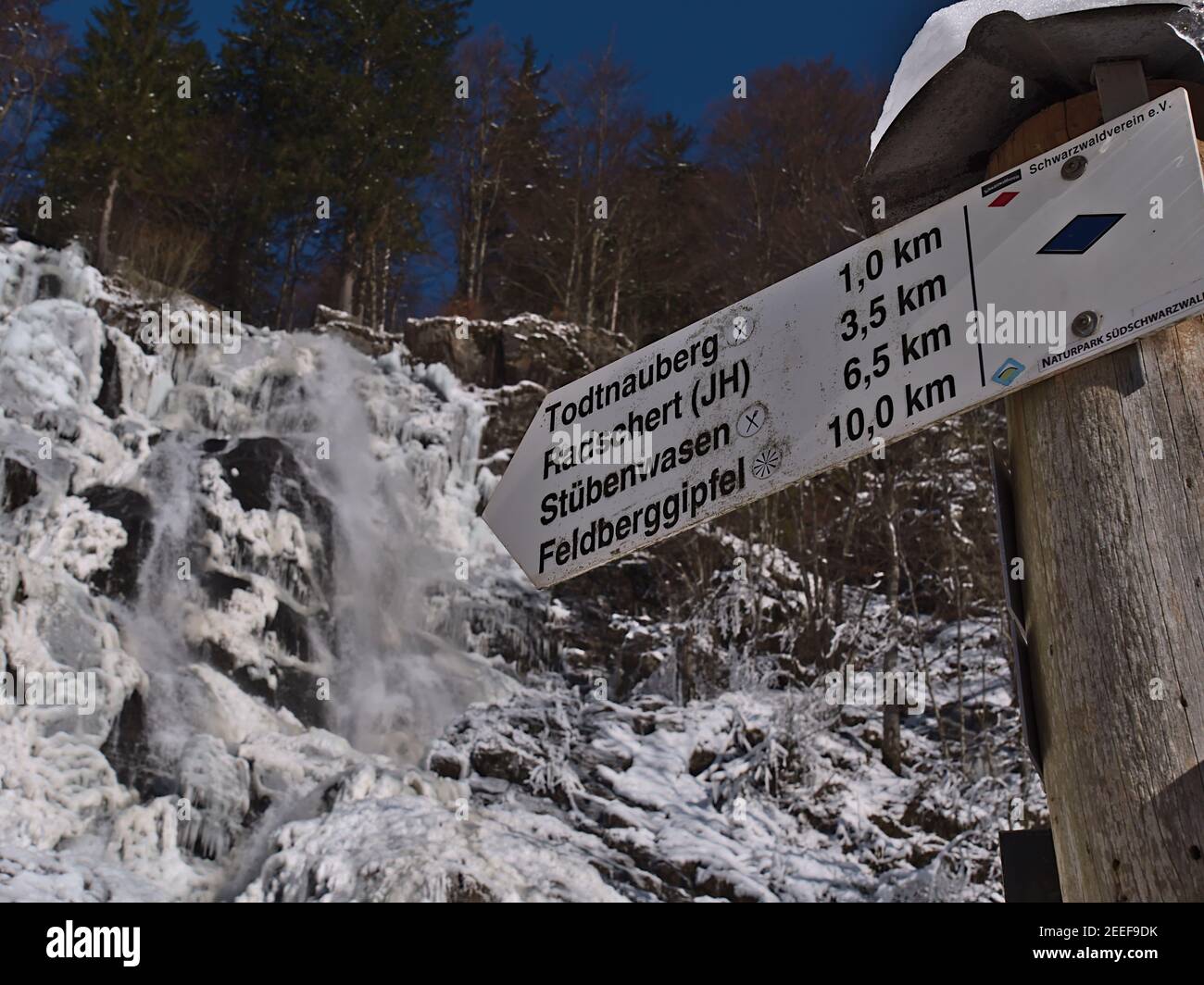 Signpost with hiking destinations and distances in front of famous waterfall Todtnauer Wasserfälle in winter with ice and snow. Focus on center. Stock Photo