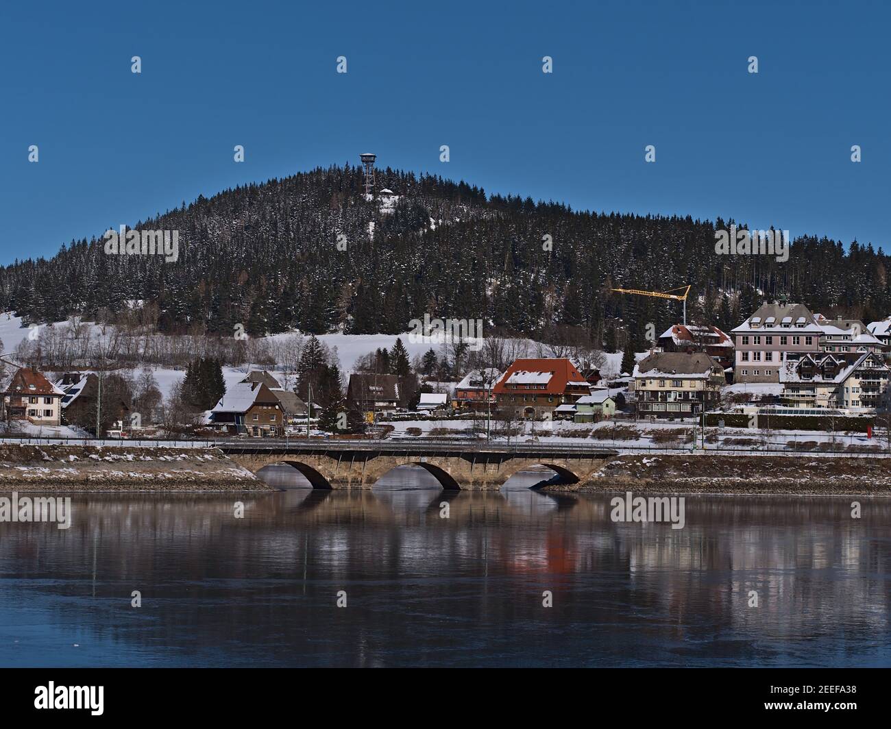Tranquil view of spa resort Schluchsee village with railway bridge reflected on the frozen lake and observation tower Riesenbühlturm on hill in winter. Stock Photo