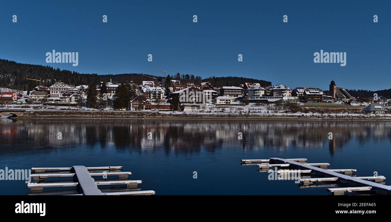 Beautiful panoramic view of small village Schluchsee located on the shore of frozen lake with snow-covered landing stages in front on sunny winter day. Stock Photo