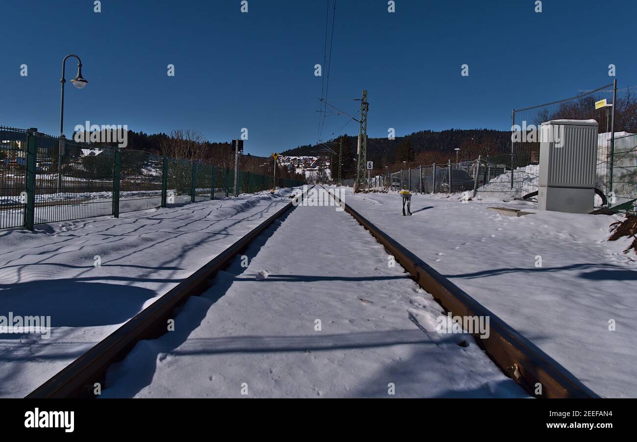 Diminishing perspective of snow-covered railroad tracks with overhead cable surrounded by fences on sunny winter day with blue sky. Stock Photo