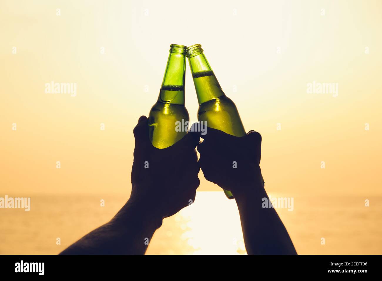 Couple hands holding beer bottles and clanging, celebrating on holiday at the beach in summer sunset Stock Photo