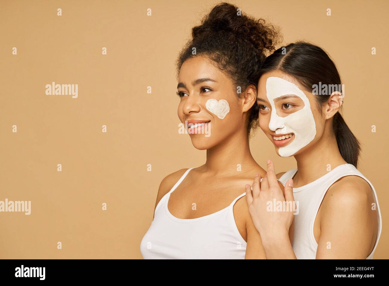 Portrait of two young women, female friends smiling aside while posing together with facial masks on isolated over beige background. Skincare, beauty concept Stock Photo