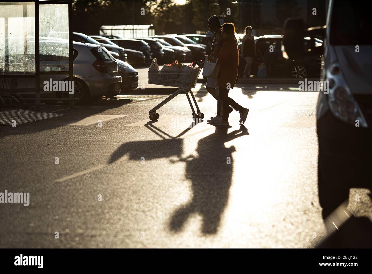 People pushing supermarket trolley out of Tesco, drawing long shadow on a sunny summer evening. Stock Photo