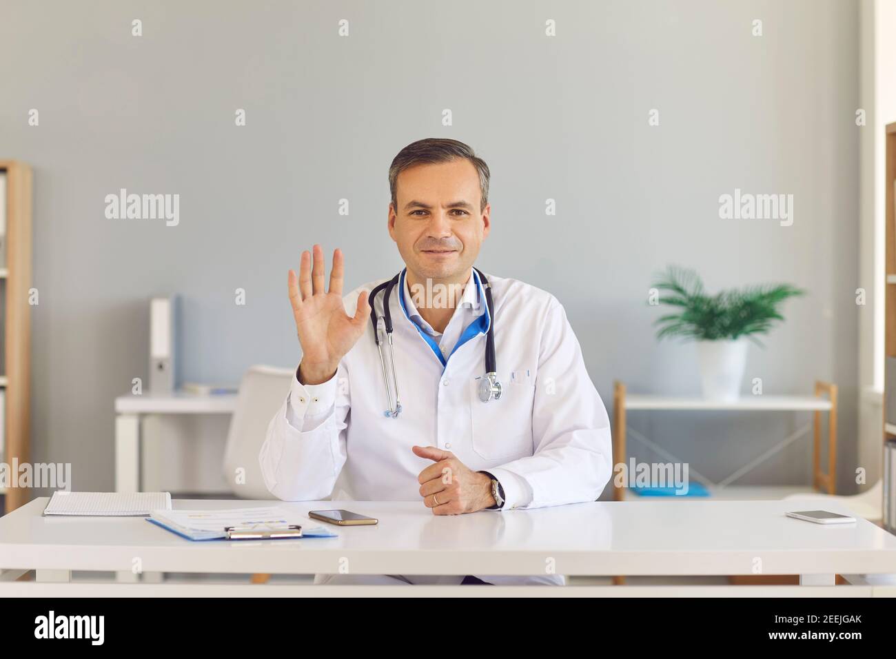Male doctor waves his hand greeting his viewers starting an online broadcast in a hospital office. Stock Photo