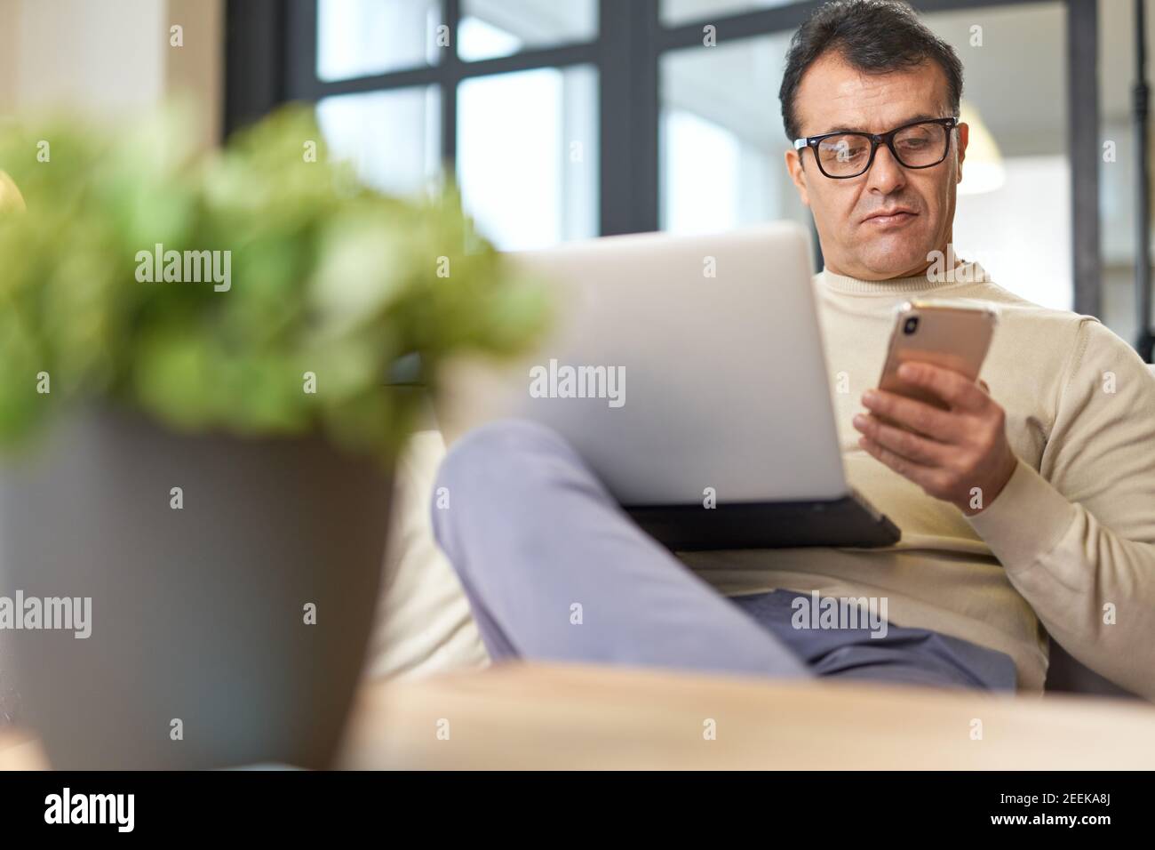 Handsome latin middle aged businessman in eyeglasses using smartphone while working on the laptop from home. Telework, business concept Stock Photo