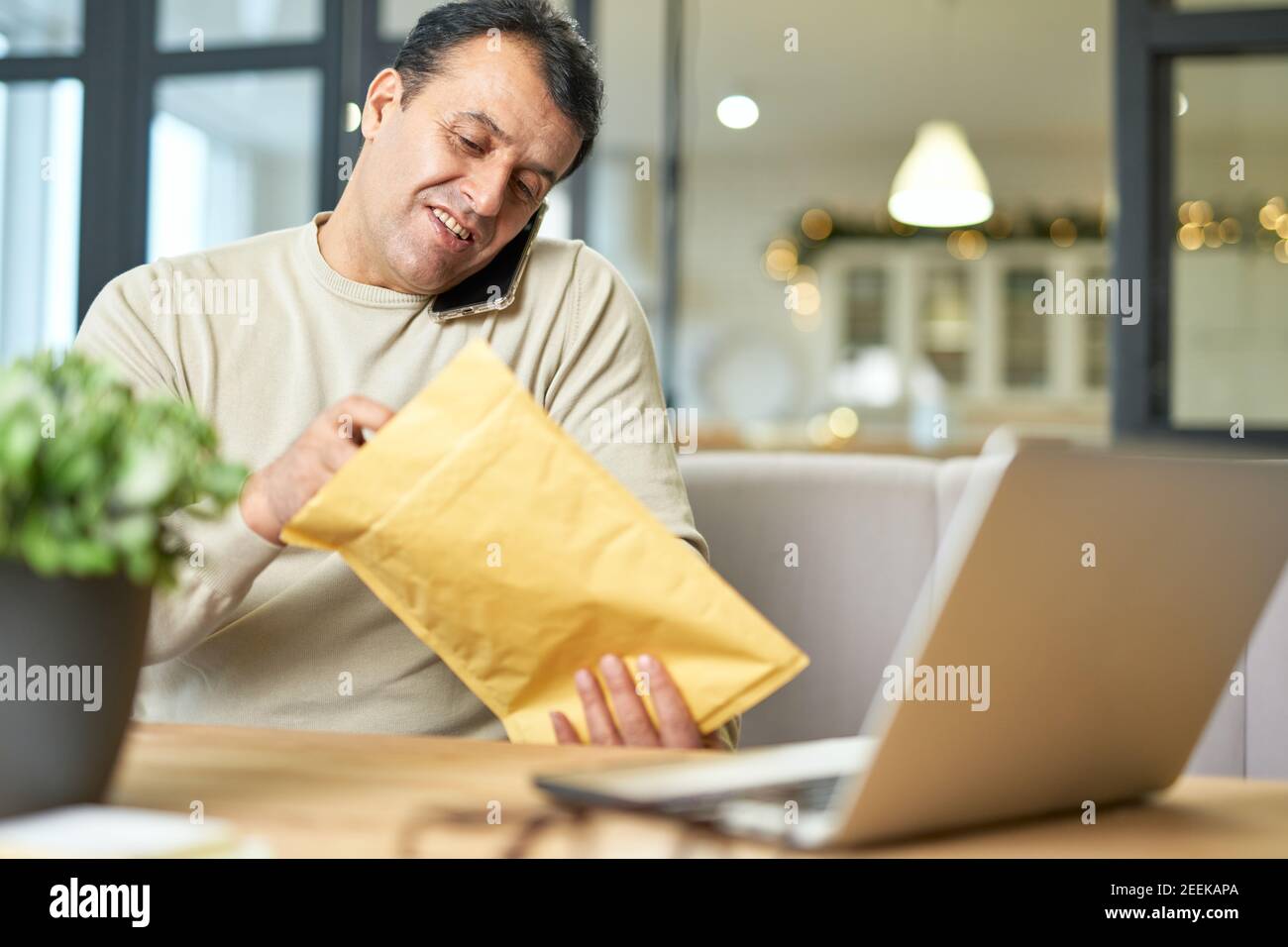 Portrait of hispanic middle aged man talking on the phone while working from home, sitting at the desk with his laptop. Focus on the man. Telework, business concept Stock Photo