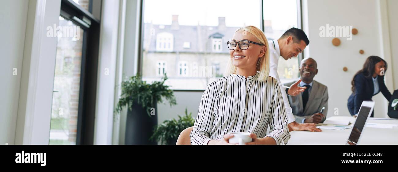 Smiling young businesswoman drinking coffee and looking deep in thought while sitting at a boardroom table with colleagues in the background Stock Photo