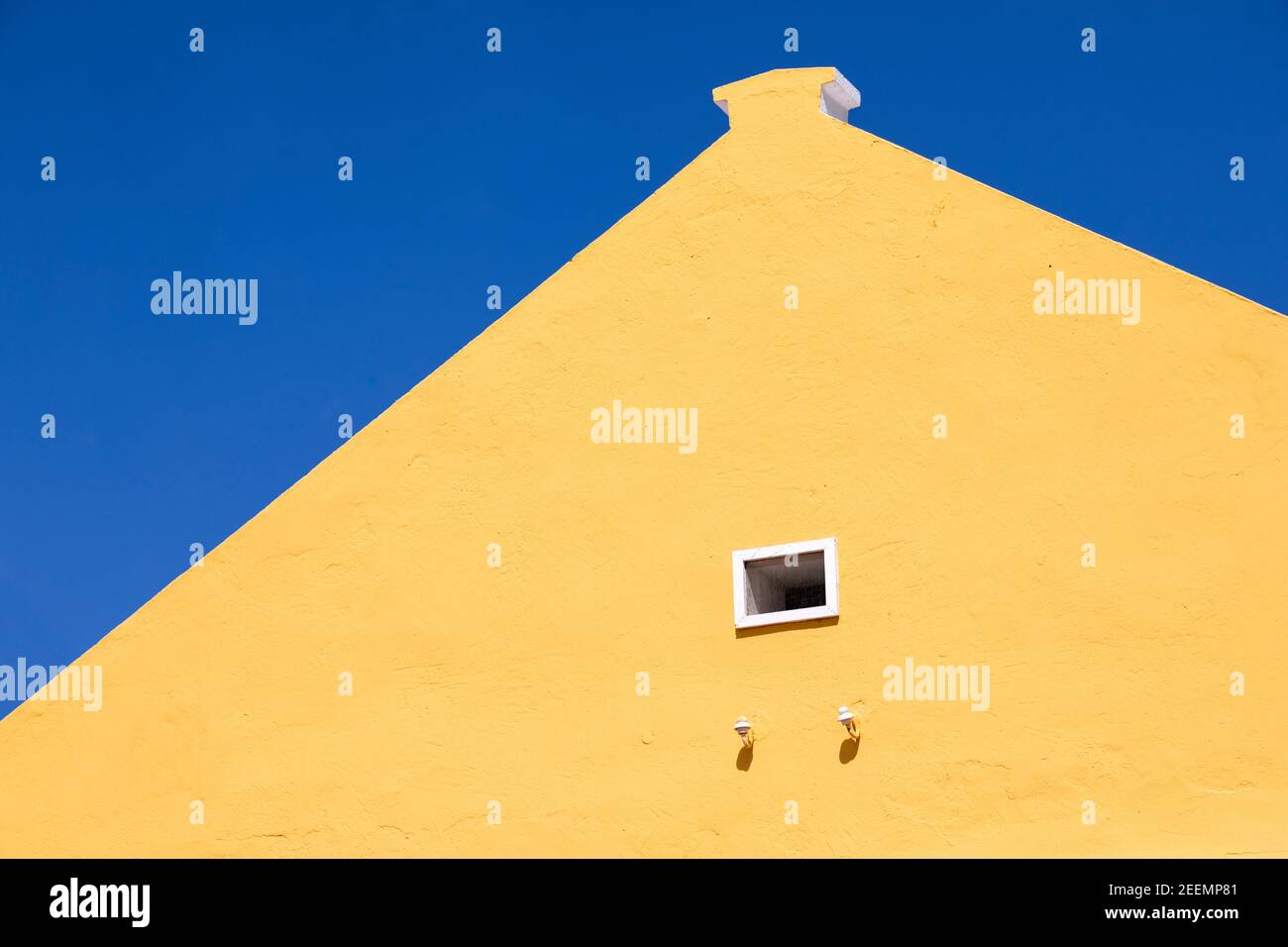 Bright yellow painted gable end of a Caribbean building with a small white framed window looking upwards against a clear deep blue sky with copy space Stock Photo