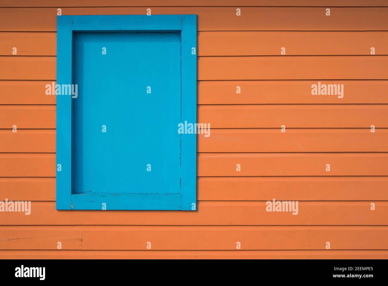 Sunlit side of an orange painted boarded wooden hut with a blocked up blue painted window with room for copy Stock Photo