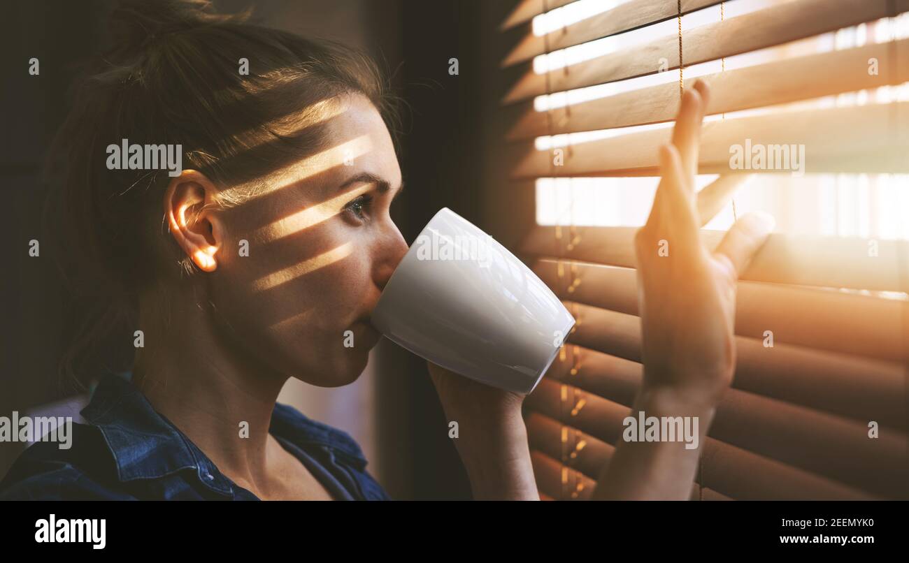 woman looking through window blinds into the sunlight and drinking coffee Stock Photo