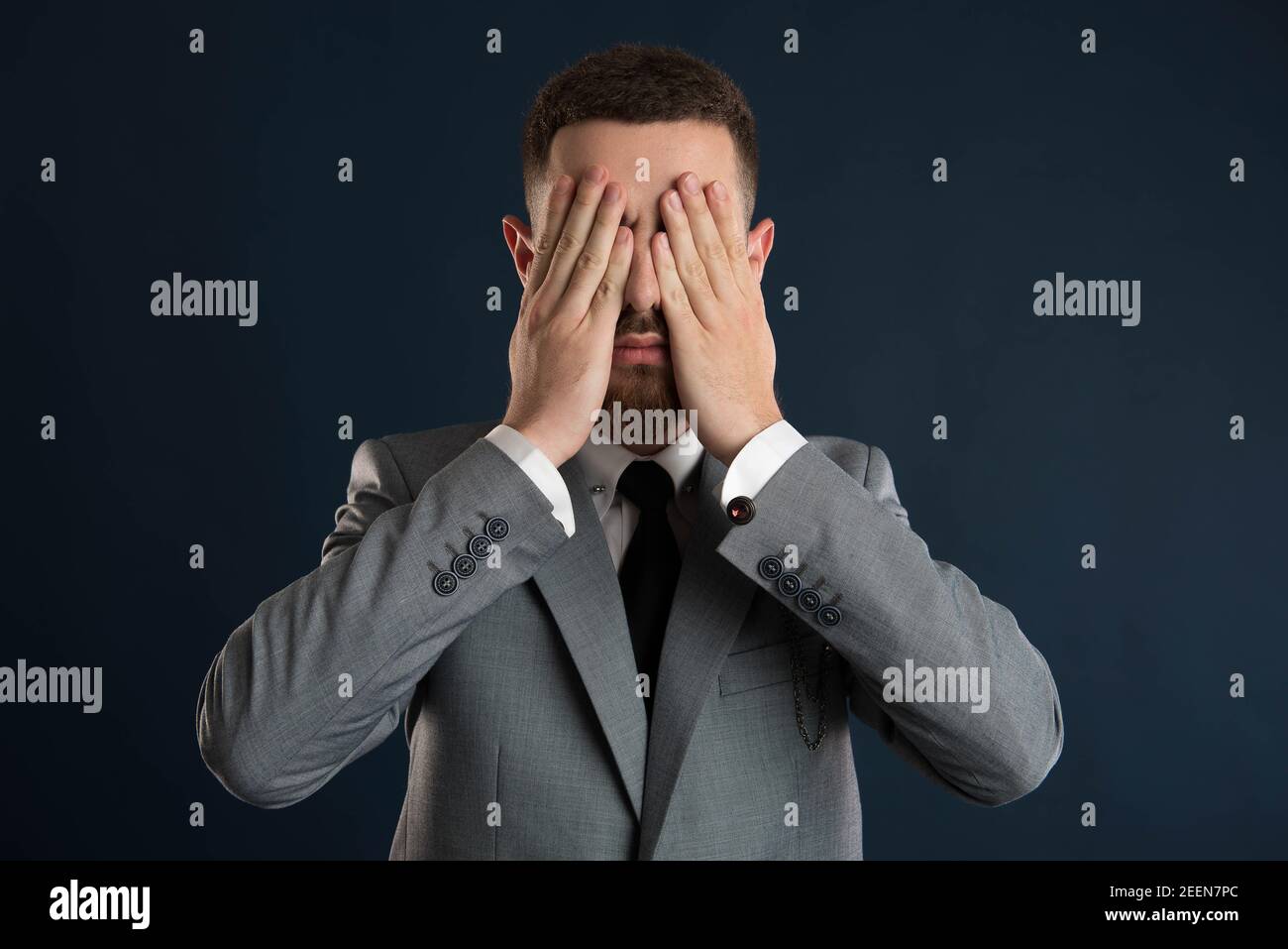 Handsome young businessman with his hands to his eyes wearing a grey suit and black tie Stock Photo