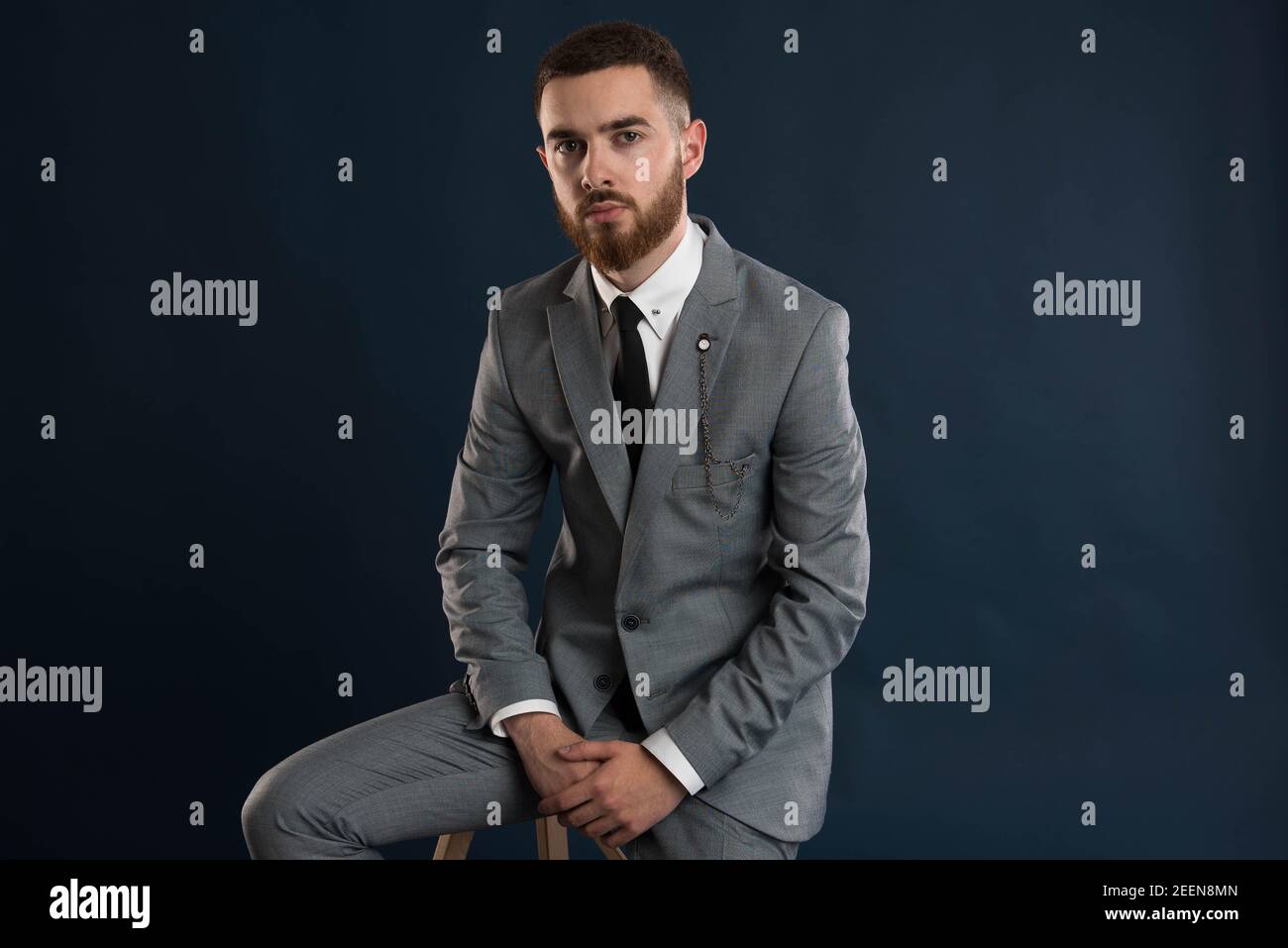 Portrait of a young gentleman sitting on a stool wearing a grey suit and black tie Stock Photo