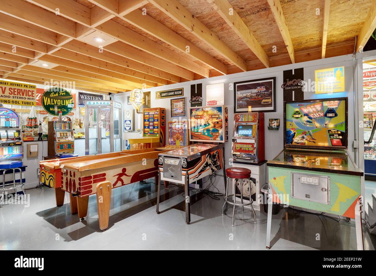 A basement arcade with various retro pinball machines and classic arcade games, old steel and tin company signs, and plenty of seating. Stock Photo