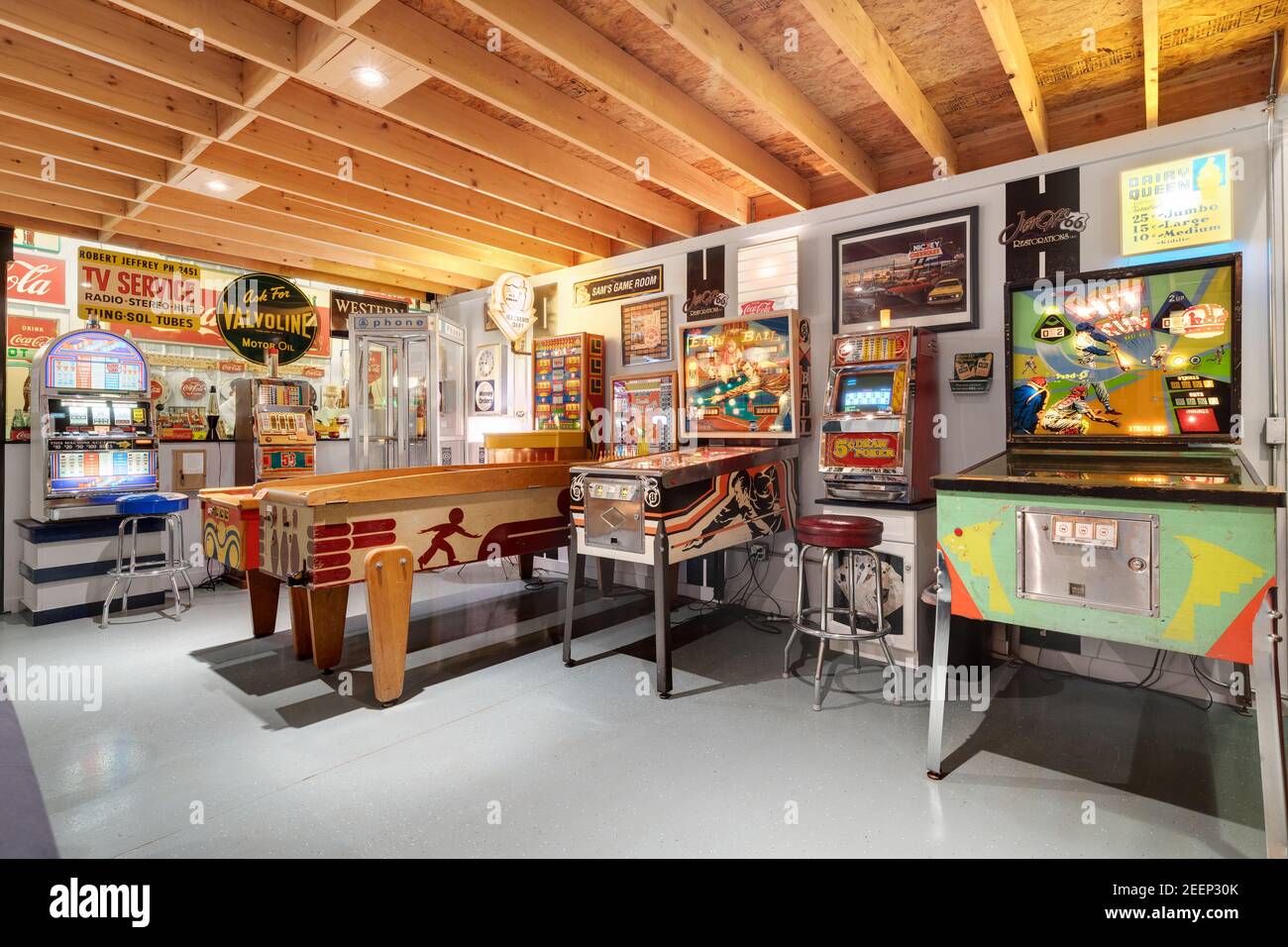 A basement arcade with various retro pinball machines and classic arcade games, old steel and tin company signs, and plenty of seating. Stock Photo