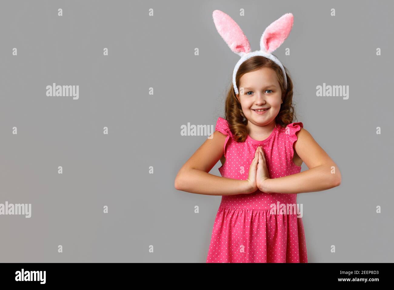Happy easter. Smiling little girl in a pink dress with polka dots on a gray background. The child in the rabbit ears folded her hands for prayer. Stock Photo