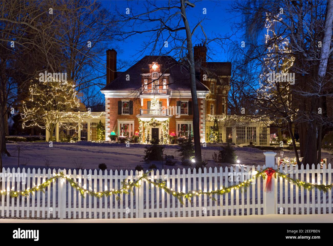 An historic 1840's brick house decorated for Christmas in a snowy winter setting, white picket fence and christmas lights at dusk Stock Photo