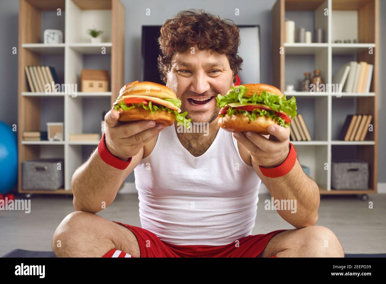 Man sportsman in sportswear sitting and holding two fresh unhealthy burgers in hands Stock Photo