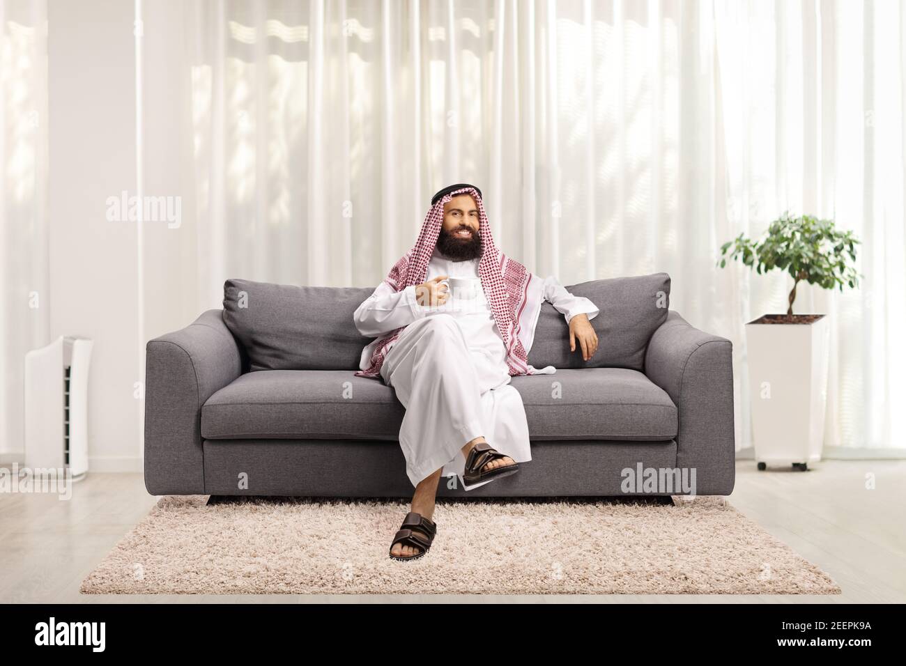 Saudi arab man sitting on a sofa at home and holding a cup of tea Stock Photo