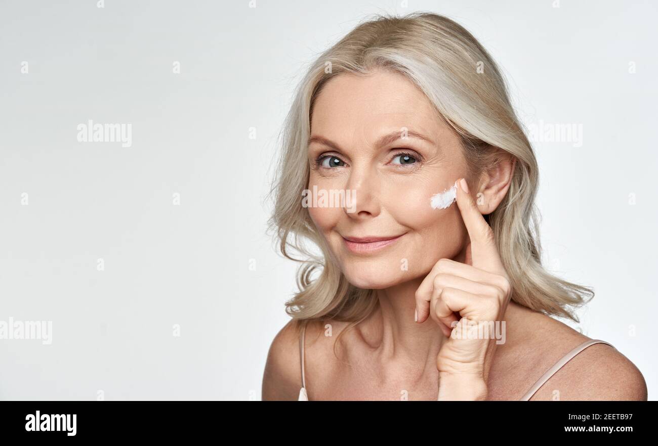 Smiling 50s middle aged mature woman applying cream on face isolated on white. Stock Photo