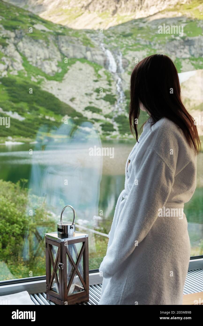 Young woman in a white bathrobe standing in front of window and enjoying the nature. SPA and wellness concept. Stock Photo