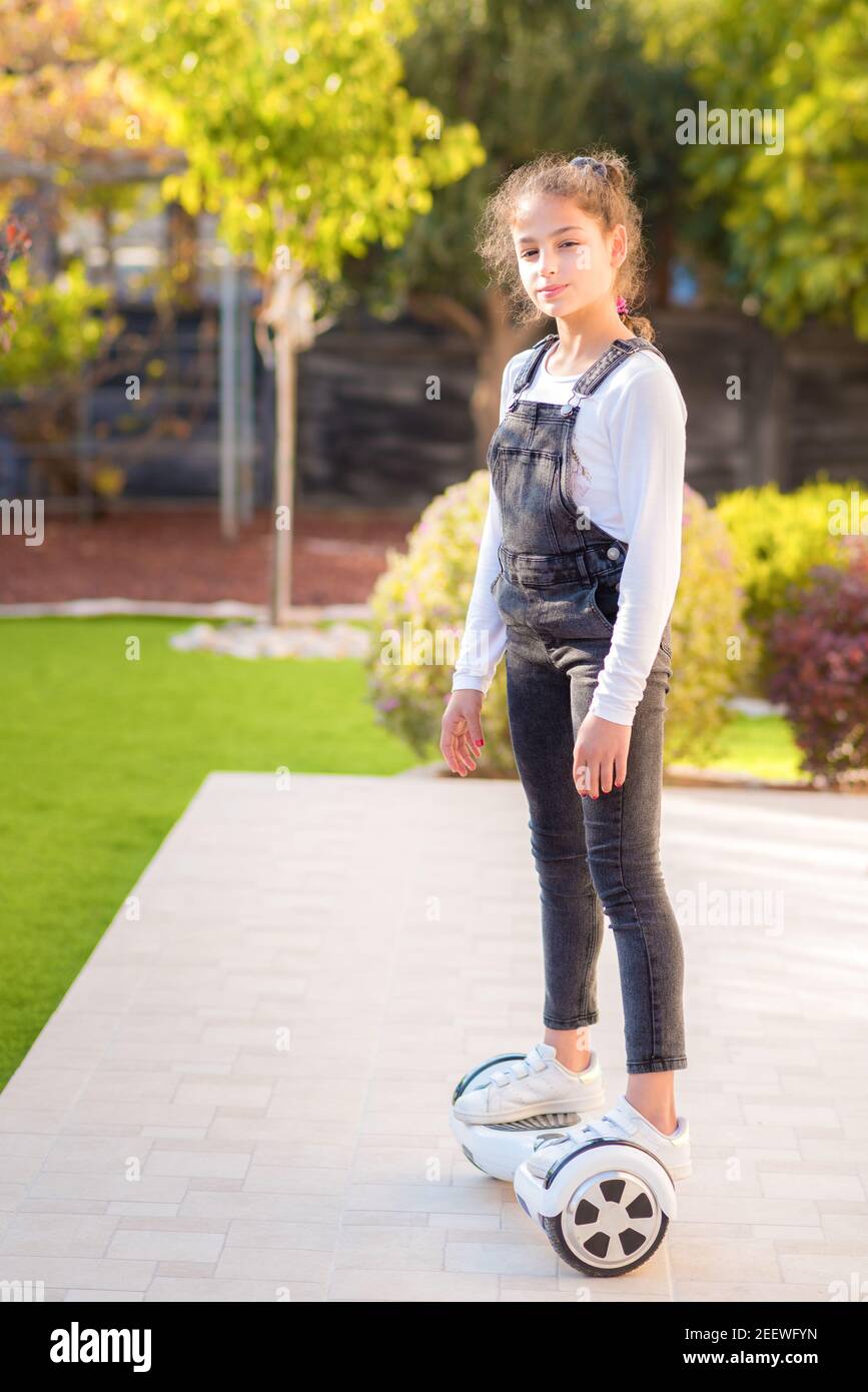 Young happy teenager girl balancing on electric hoverboard at the sunny park, sunny day. Child using a self-balancing two-wheeled board.Beautiful kid using Hover Board. Side view. Stock Photo