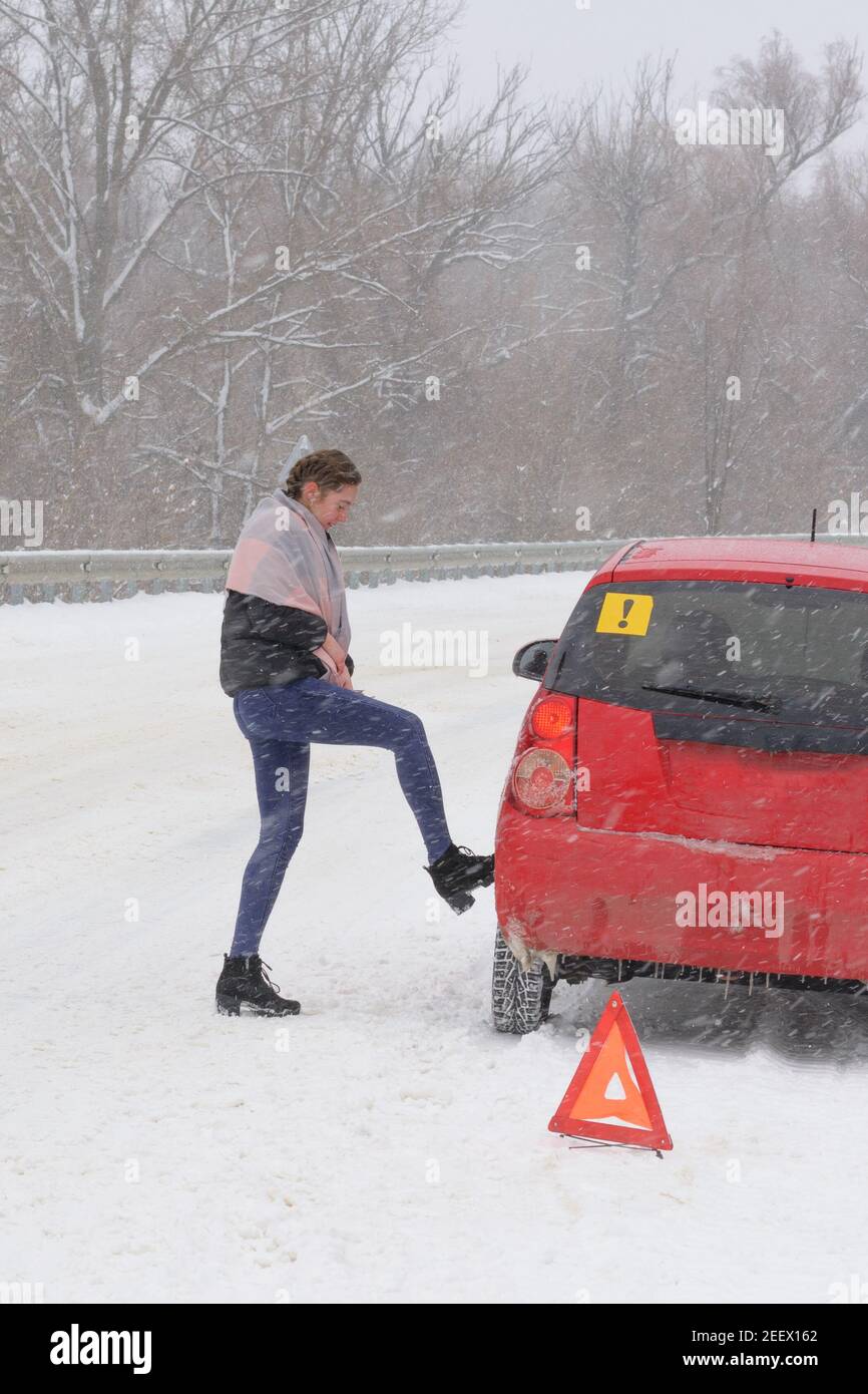 Young woman driver checks tire of  car on road in  blizzard and bad weather. Triangular accident warning sign stands on the roadway. Winter driving. C Stock Photo