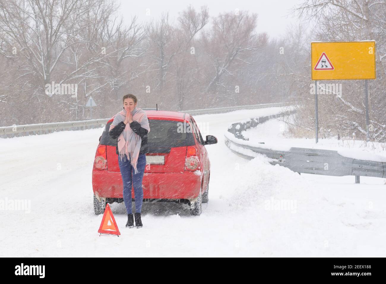 Confused woman driver near broken car on slippery road. Snow and blizzard, winter driving hazard. Stock Photo