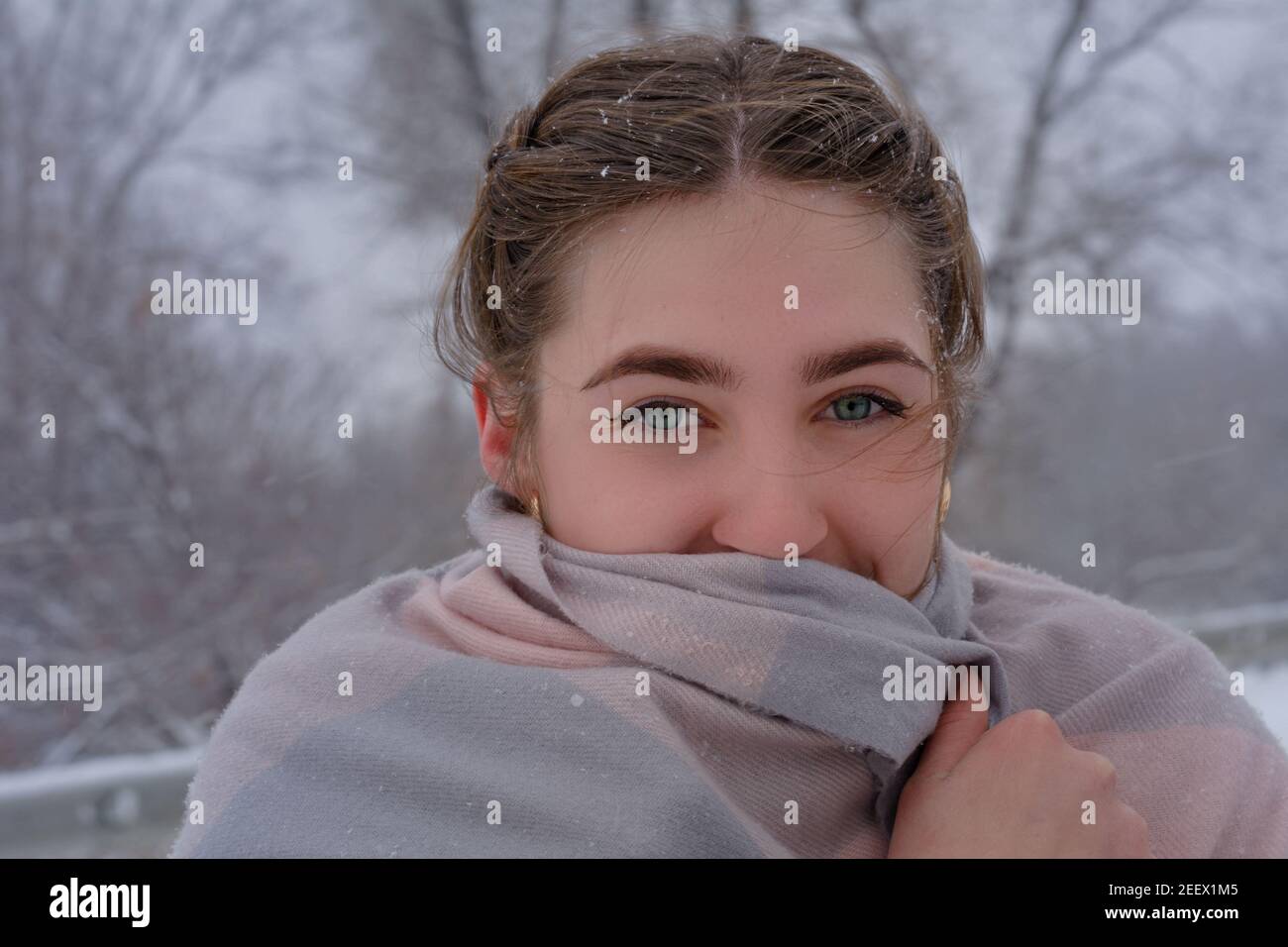 Young woman with beautiful green eyes wears scarf in winter outdoors. Lady froze snow, snowflakes in her hair. Close-up. Stock Photo