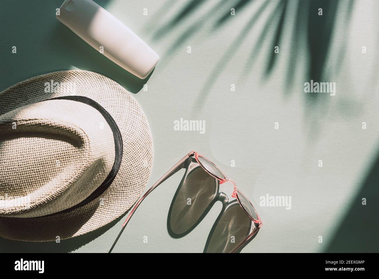 Sun hat, glasses and spf cream or lotion unbranded package on light blue background with palm tree leaf shadow, top view, flat lay. Summer vacation co Stock Photo