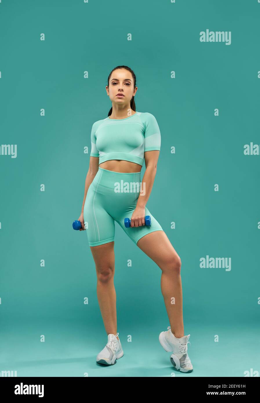 Beautiful fit lady with dumbbells looking at camera with serious expression. Isolated on blue background Stock Photo