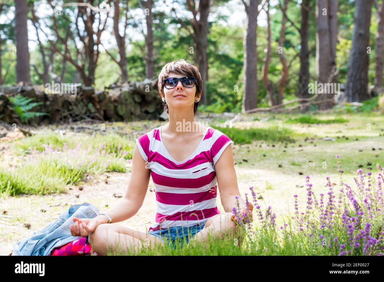 A woman meditating after hiking in the summer forest. Enjoy the sun on the meadow with flowers. Unity with nature, digital detox, personal fulfillment Stock Photo