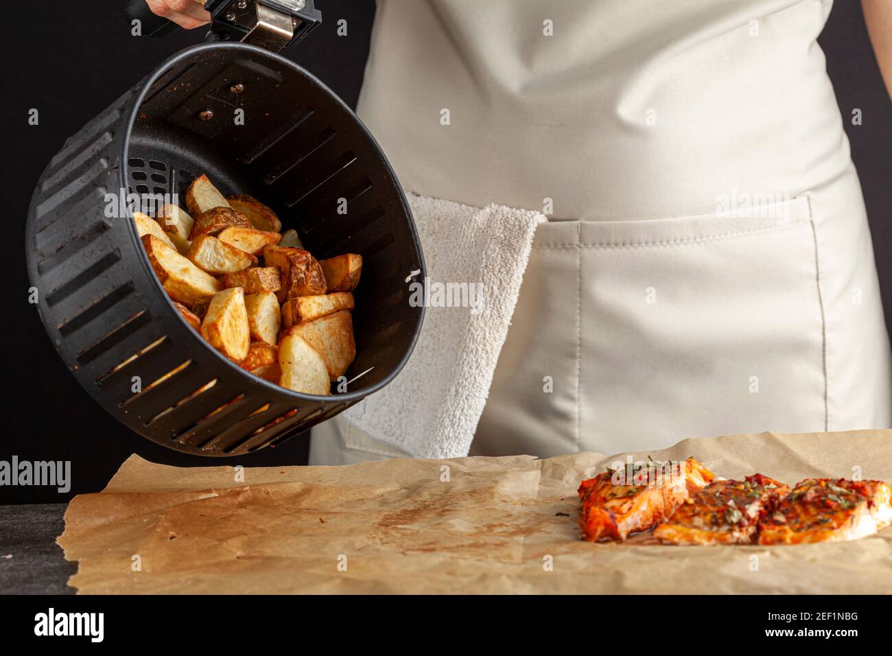 A caucasian woman chef is dumping fresh made fried potato chunks on a piece of baking paper which has three oven roasted marinated salmon fillet with Stock Photo