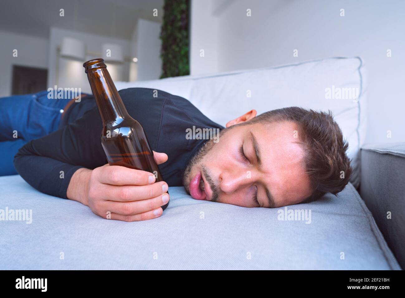 Drunk young man with open mouth and bottle of beer sleeping on the sofa at home. Stock Photo