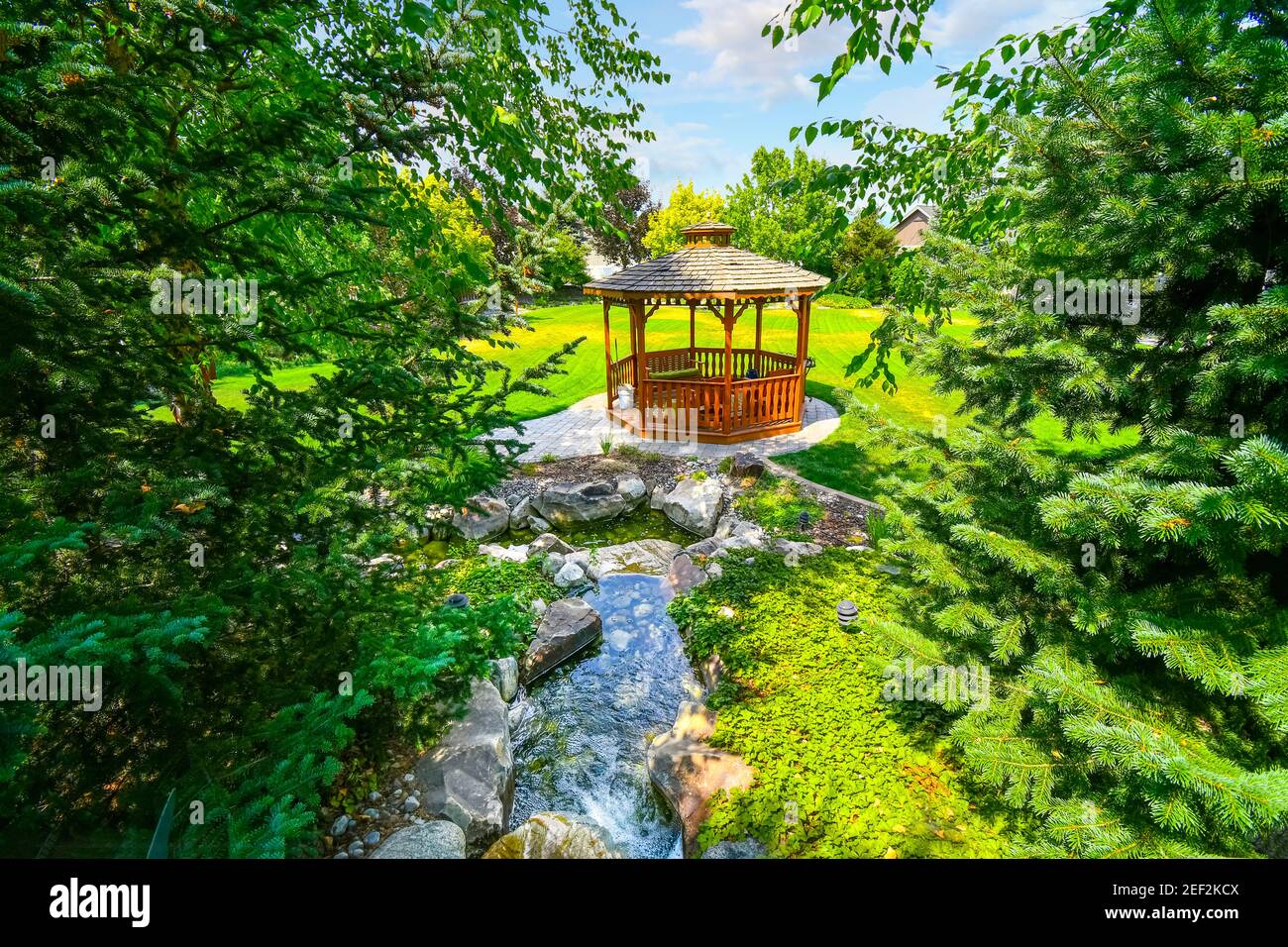 A small stream flows towards a wooden gazebo in a landscaped garden. Stock Photo