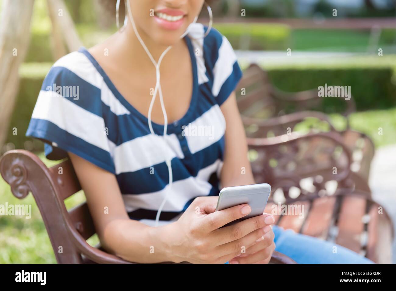 Young black woman sitting on the bench in the garden and listening to music from smartphone Stock Photo