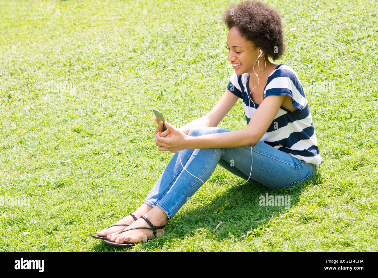 Casual afro woman sitting on the grass and listening to music from smartphone Stock Photo