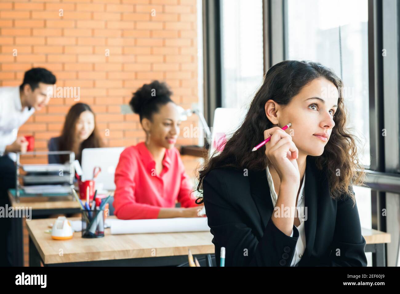 Businesswoman sitting in the office, looking up and thinking with pencil at her cheek Stock Photo