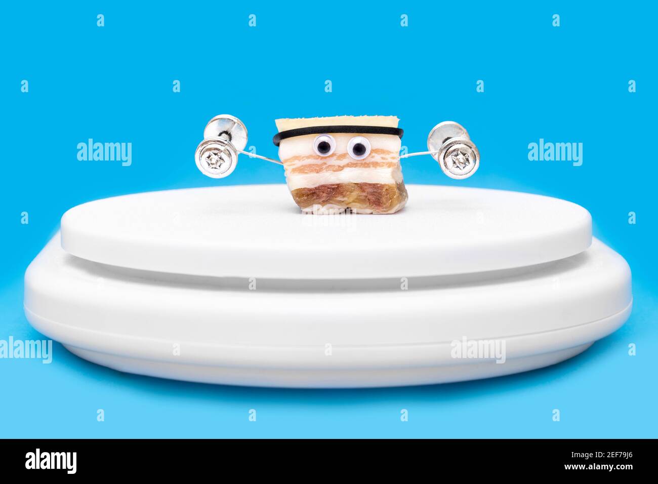Close-up of a funny character made of salted pork fat lifting dumbbells standing on kitchen scales against a light blue background. Creative fat burni Stock Photo