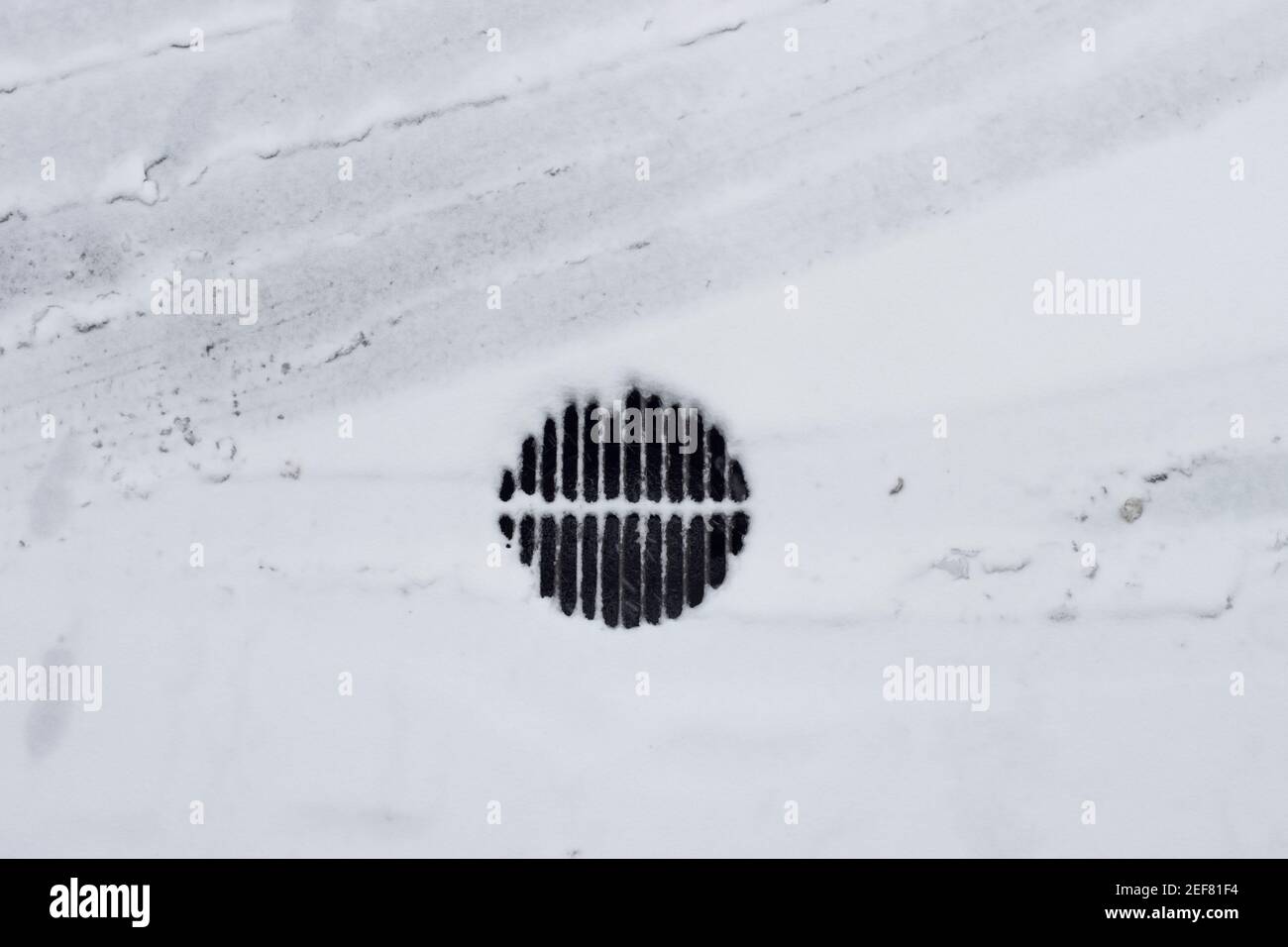 Top view of frozen and snow-covered storm drainage grate, car tracks and footprints in the snow. Random elements on the street in winter. Stock Photo