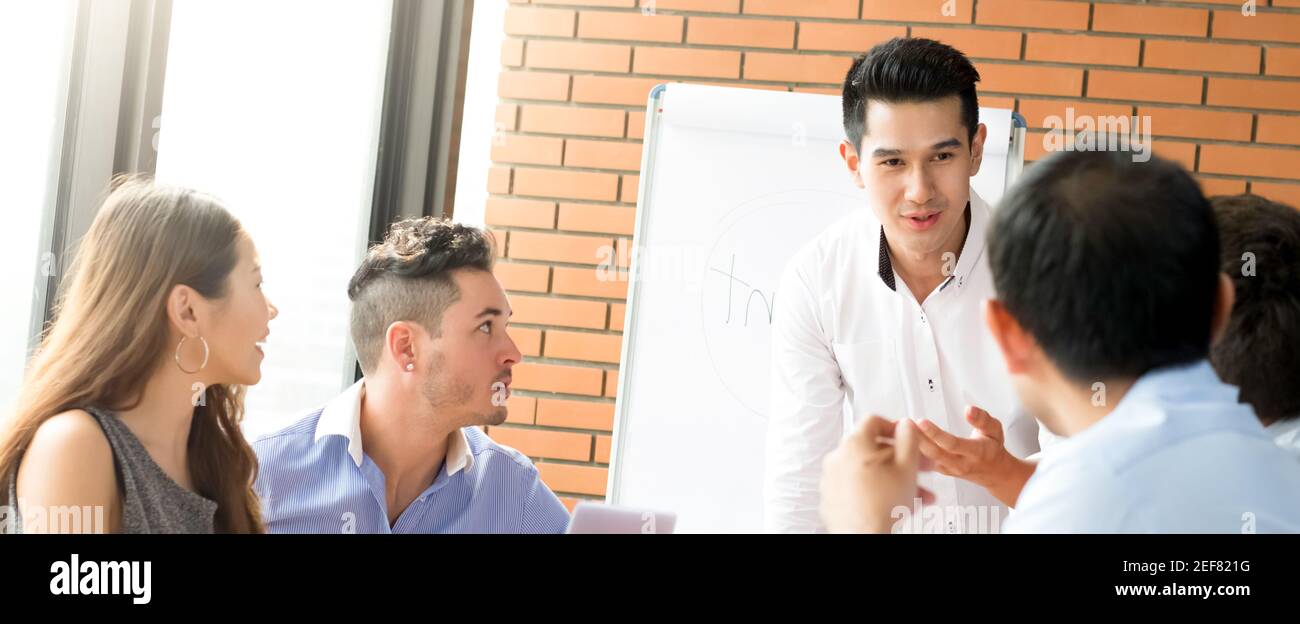 Young casual Asian businessman as a meeting leader presenting his work to mixed race colleagues, panoramic banner Stock Photo