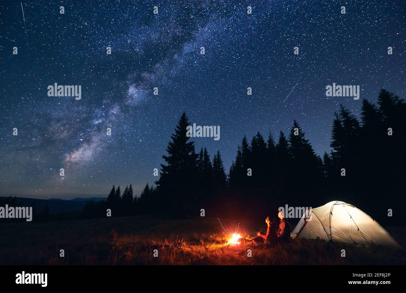 Young Couple Hikers Sitting Near Bright Burning Bonfire And Illuminated Tourist Tent Enjoying Camping Night Together Under Dark Sky Full Of Shiny Stars And Bright Milky Way Warm Summer Evening Stock Photo