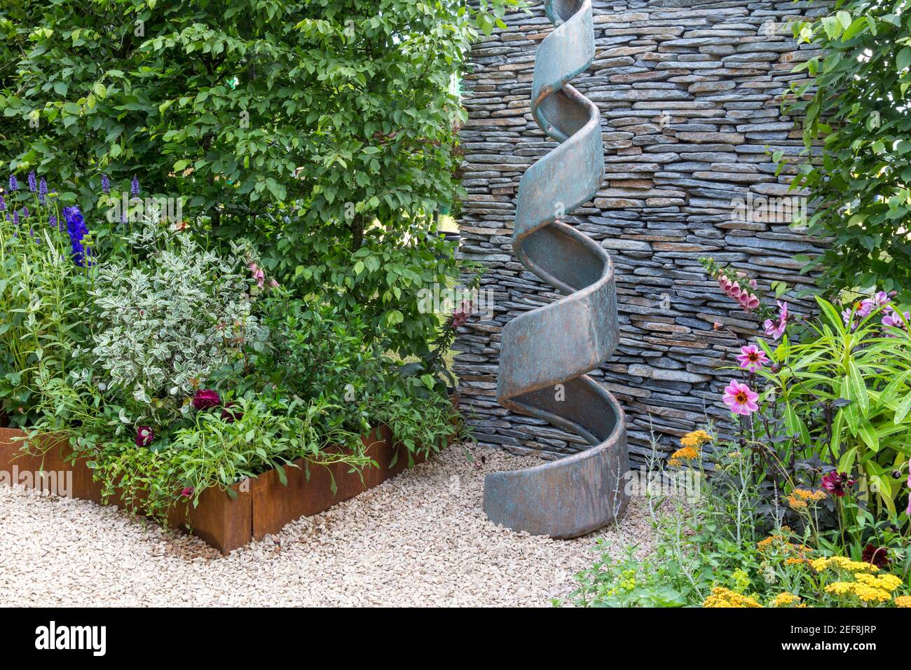 Small English courtyard cottage garden with spiral sculpture - dry stone wall - hedge - gravel path with garden flower borders England UK Stock Photo