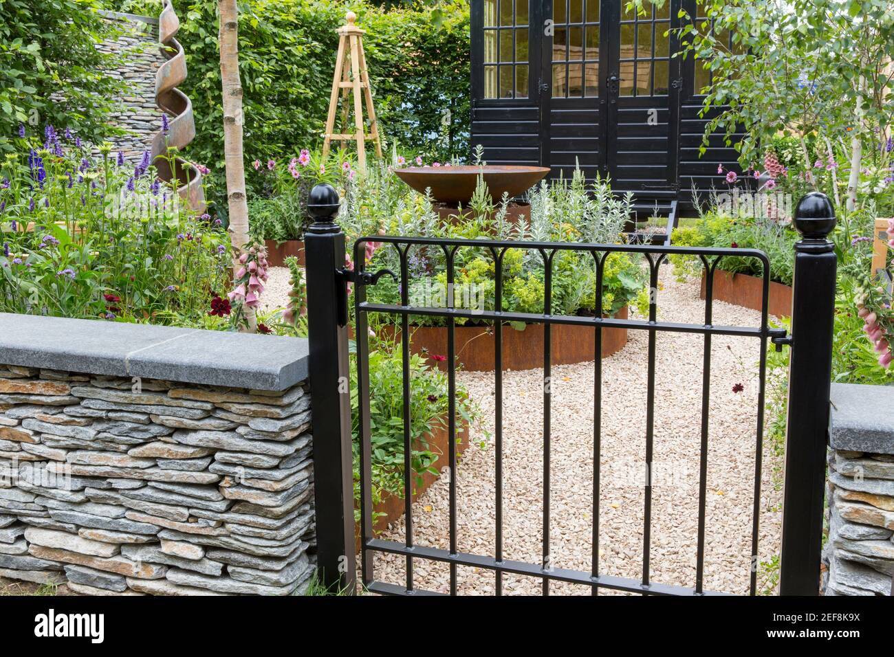 English cottage country garden A painted black metal garden gate - dry stone wall with gravel path and flower beds garden border England UK Stock Photo