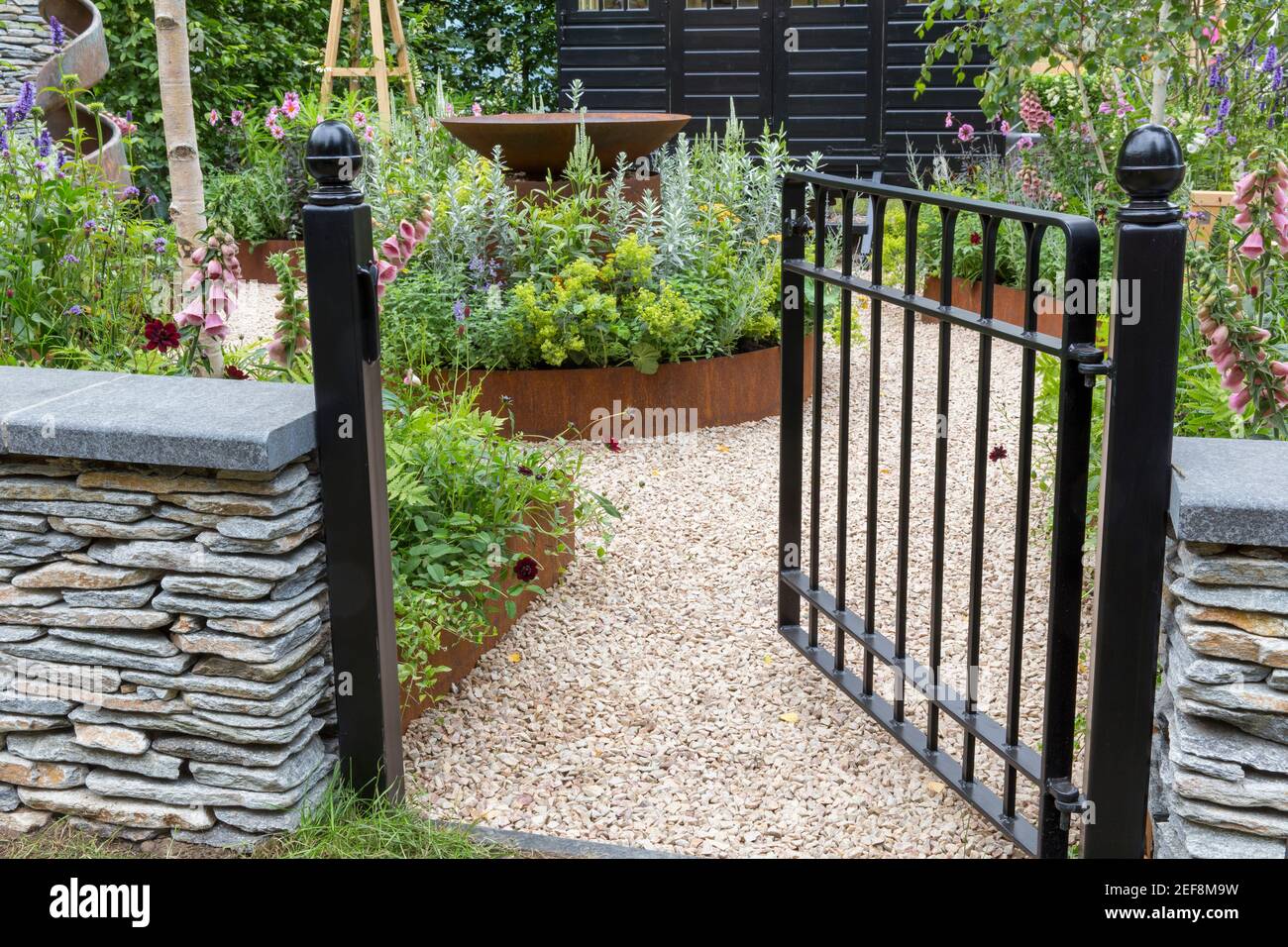 English country cottage garden A painted black metal garden gate with dry stone wall view of gravel path and flower beds borders England UK Stock Photo