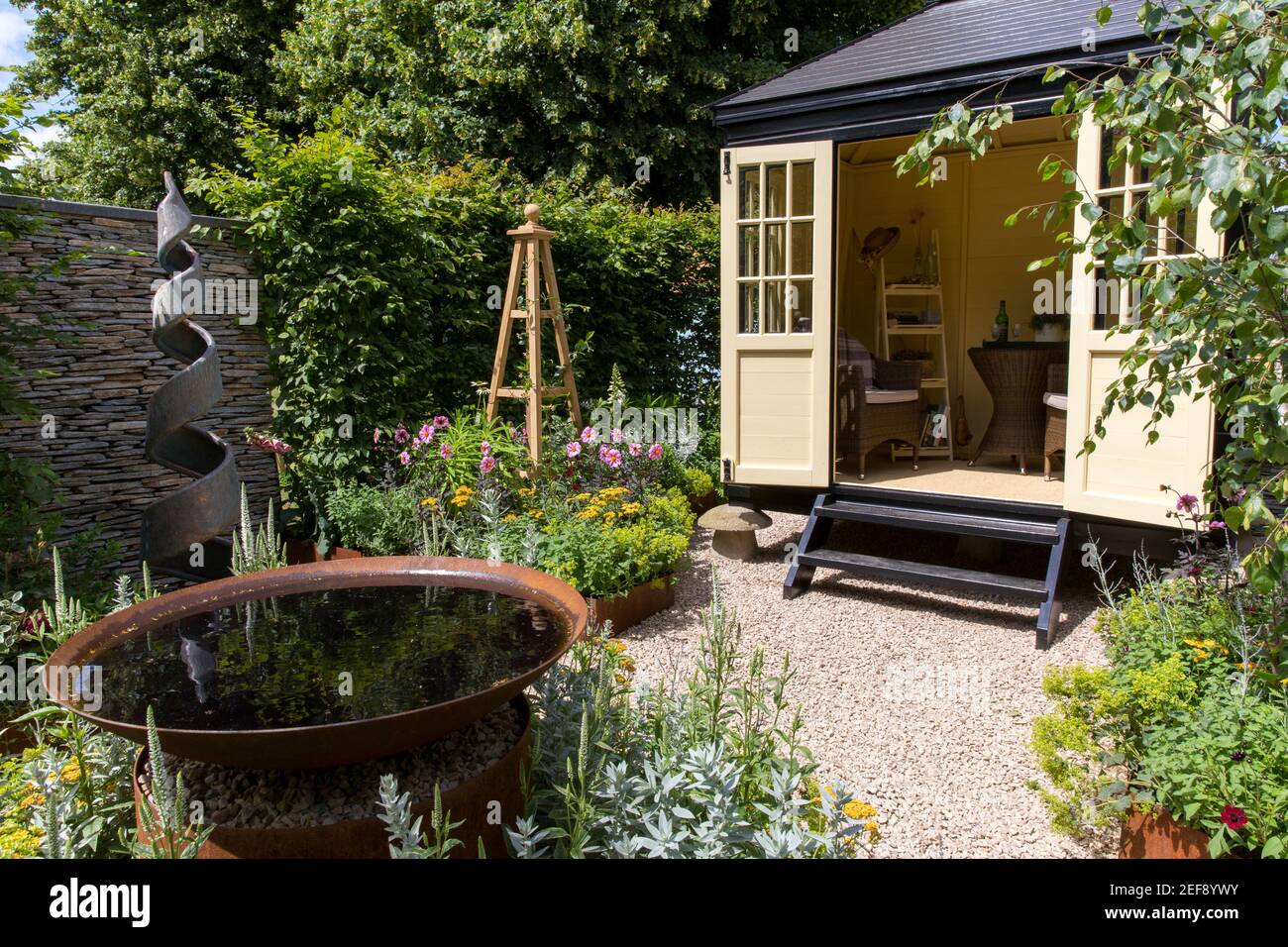 A small English cottage garden with Summerhouse Shepherds Hut in gravel garden - working from home office - water feature spiral sculpture obelisk UK Stock Photo