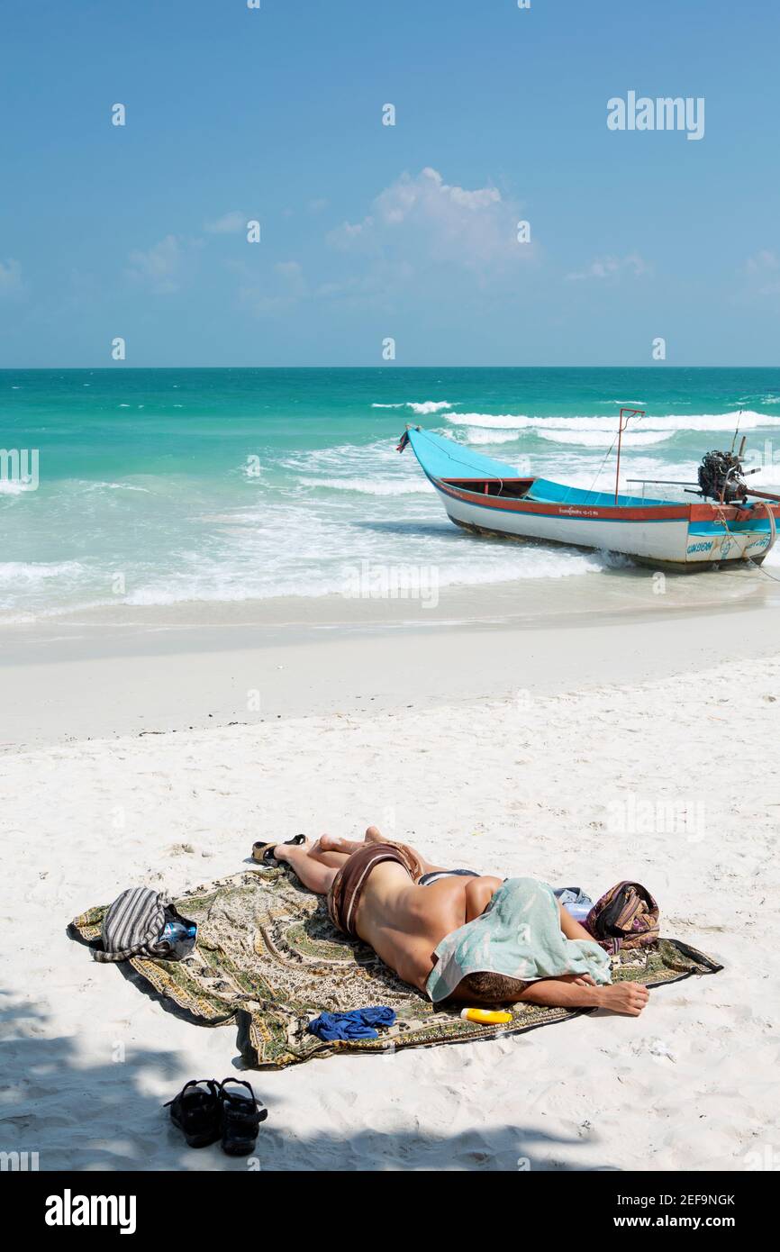 A backpacker asleep on the beach in the sun, copy space, thai boat, hungover, Haad Rin party beach, Koh Phangan island, Thailand Stock Photo