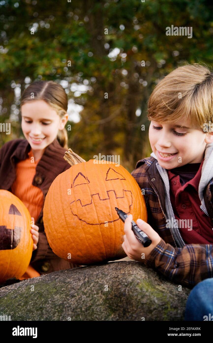 Boy drawing a human face on a pumpkin with his sister sitting behind him Stock Photo