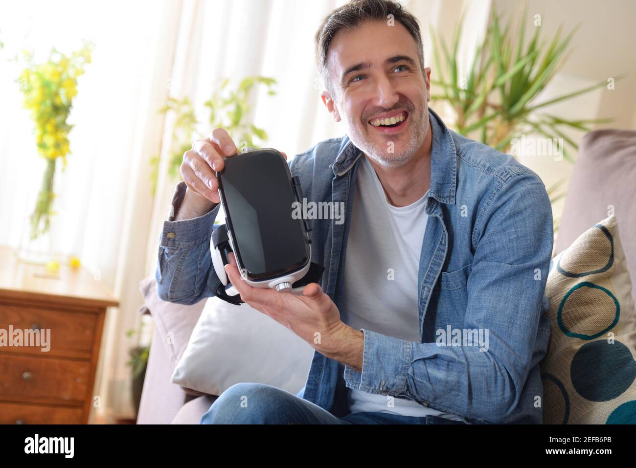 Smiling man showing virtual reality glasses sitting on a sofa in the living room at home Stock Photo