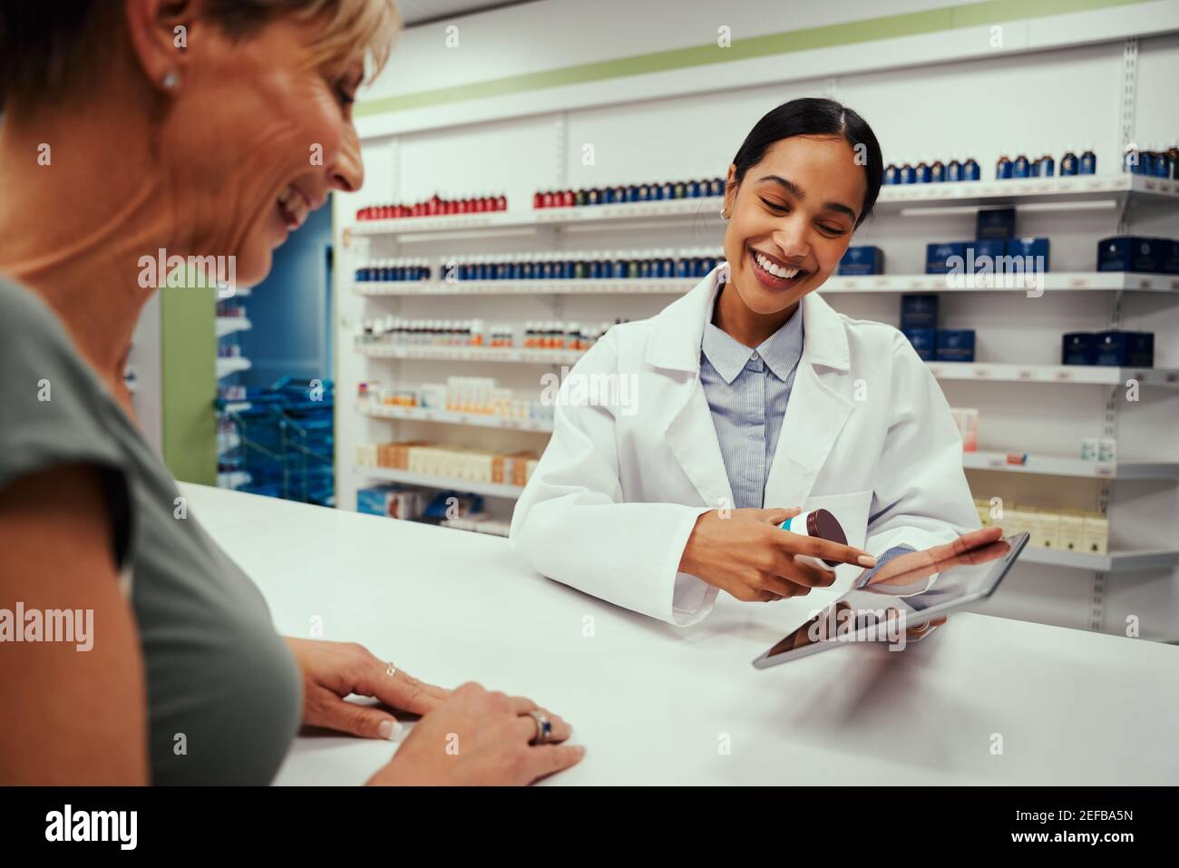 Cheerful young woman wearing labcoat working in pharmacy showing screen to female customer on digital tablet Stock Photo