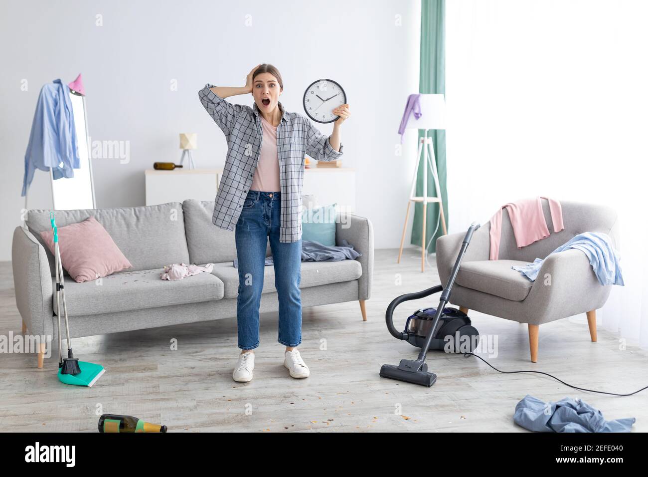 Shocked young lady with clock grabbing her head in terror, standing in messy room after party, too late to clean apartment before return of her parent Stock Photo